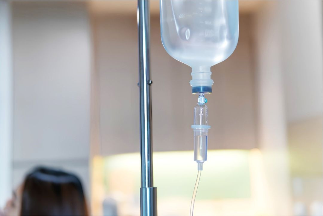 Close-up focado em um equipamento de gotejamento de soro hospitalar transparente, preso a um suporte metálico. O frasco com o líquido está pendurado de cabeça para baixo, conectado a um tubo fino de infusão intravenosa que desce em direção ao paciente. Ao fundo, com desfoque de imagem, nota-se o ambiente de uma clínica de saúde e parte do cabelo de uma pessoa deitada.