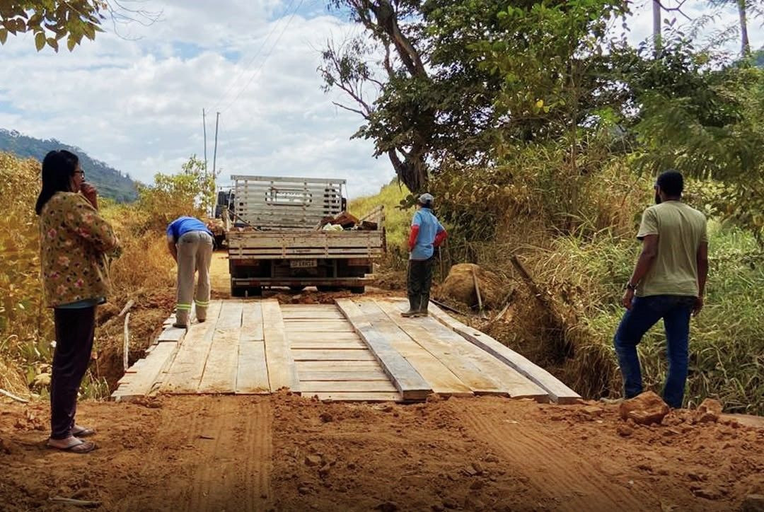 Fotografia horizontal em plano médio mostrando o andamento da obra na ponte de madeira de Terra Corrida, em Iúna. Um caminhão de carroceria aberta está estacionado sobre a estrutura de madeira recém-instalada. Quatro pessoas aparecem na imagem: uma mulher à esquerda observando, um funcionário de calça cinza e blusa azul curvado próximo à lateral do caminhão, um homem de camisa azul clara e boné no centro e outro homem de camiseta verde e calça jeans à direita, de costas. O entorno é composto por terra batida vermelha e vegetação nativa densa sob um céu parcialmente nublado.