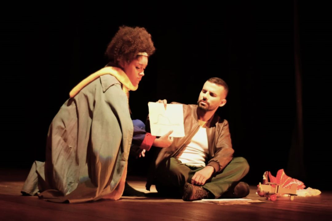 Cena de uma peça de teatro em um palco com fundo escuro. À esquerda, uma pessoa de casaco azul e cachecol amarelo, agachada, mostra um papel com um desenho para outra pessoa sentada à direita. A pessoa sentada veste uma jaqueta de couro e segura o papel. Ao lado delas, objetos de cena, incluindo o que parece ser um avião de brinquedo.