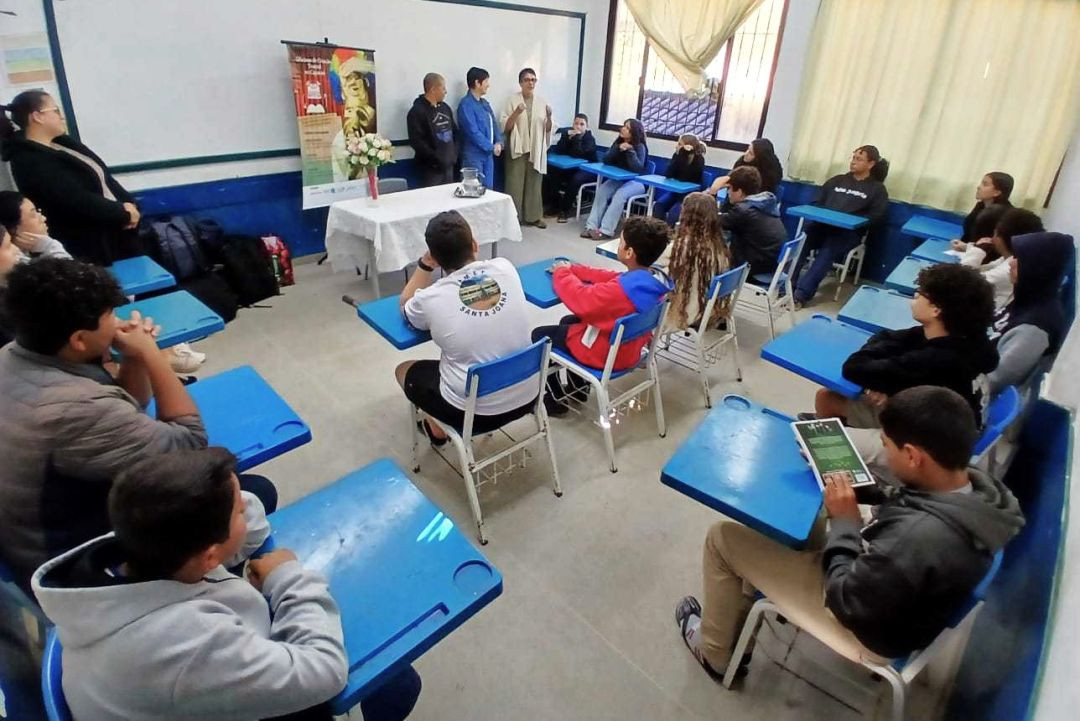 Fotografia em plano geral de uma sala de aula com paredes brancas e barrado azul. Cerca de quinze estudantes adolescentes estão sentados em carteiras escolares azuis dispostas em formato de 