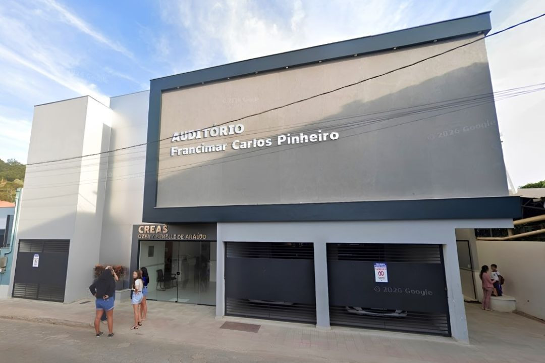 Fotografia diurna em plano médio mostrando a fachada moderna de um prédio de dois pavimentos em Dores do Rio Preto. A parte superior, de cor cinza escuro com moldura preta, exibe a inscrição em letras brancas 