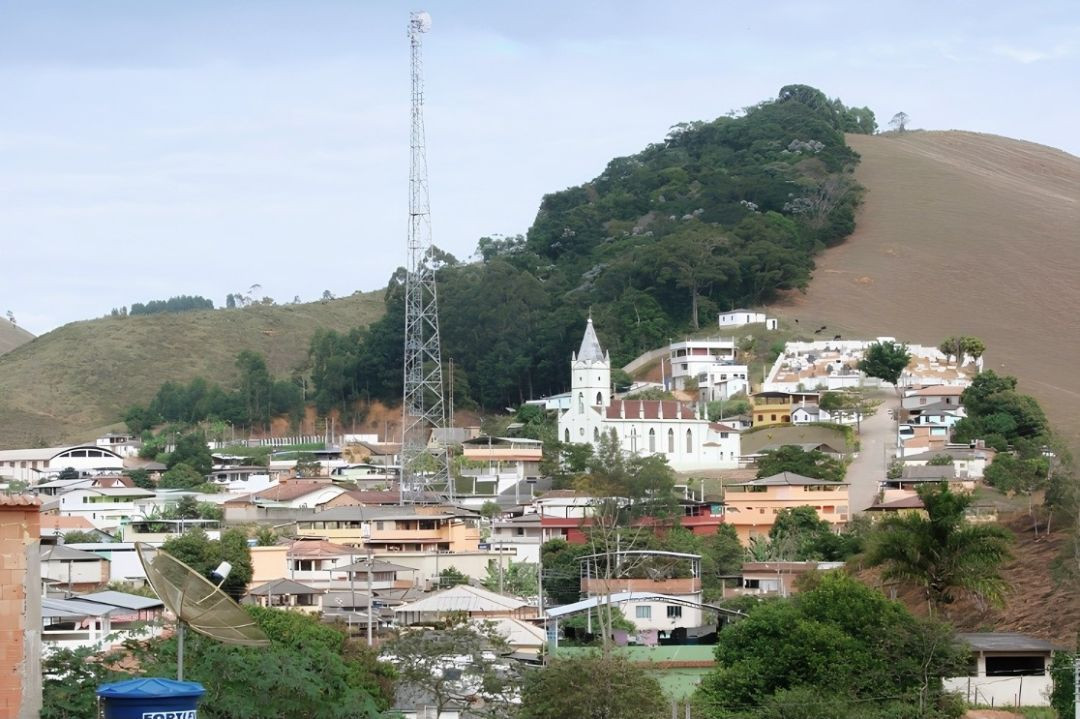 Fotografia em plano aberto mostrando a cidade de Divino de São Lourenço, no Espírito Santo. Em primeiro plano, telhados de residências, uma antena parabólica azul e uma torre de transmissão metálica alta e fina. Ao fundo, destaca-se uma igreja branca com torre pontiaguda de estilo clássico, cercada por árvores verdes e uma colina de terra batida sob um céu parcialmente nublado.