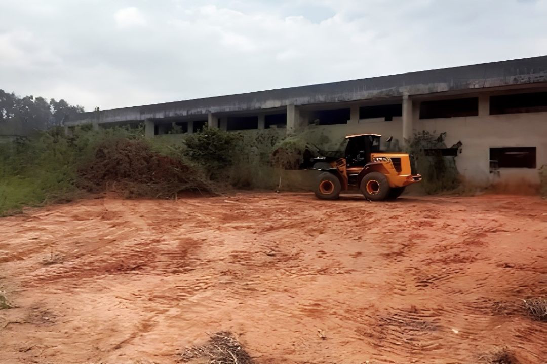 Fotografia colorida horizontal em ângulo frontal mostrando um trator carregador de cor amarela operando em um terreno de terra batida avermelhada. Ao fundo, destaca-se a estrutura de concreto de um prédio de dois pavimentos com aberturas para janelas sem vidros, cercado por vegetação rasteira e árvores sob um céu nublado.