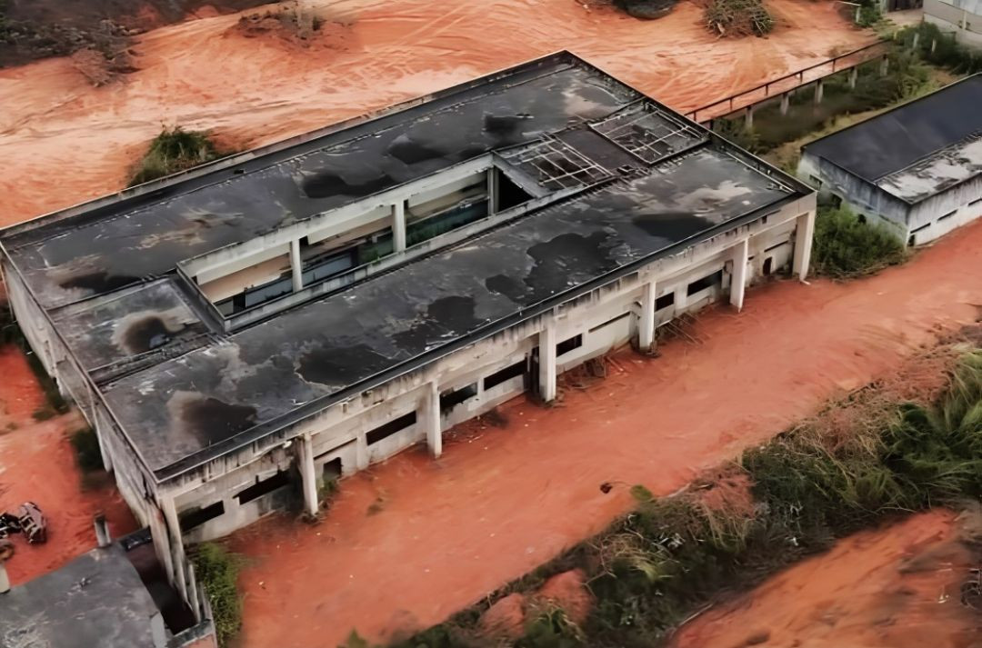 Fotografia aérea colorida tirada de um ângulo superior diagonal, mostrando a cobertura e as paredes de concreto de uma grande edificação inacabada com formato retangular vazado no centro. O topo da estrutura apresenta manchas escuras causadas pela ação do tempo, e o entorno é composto por solo de terra batida vermelha, caminhos de acesso e vegetação rasteira nas laterais.