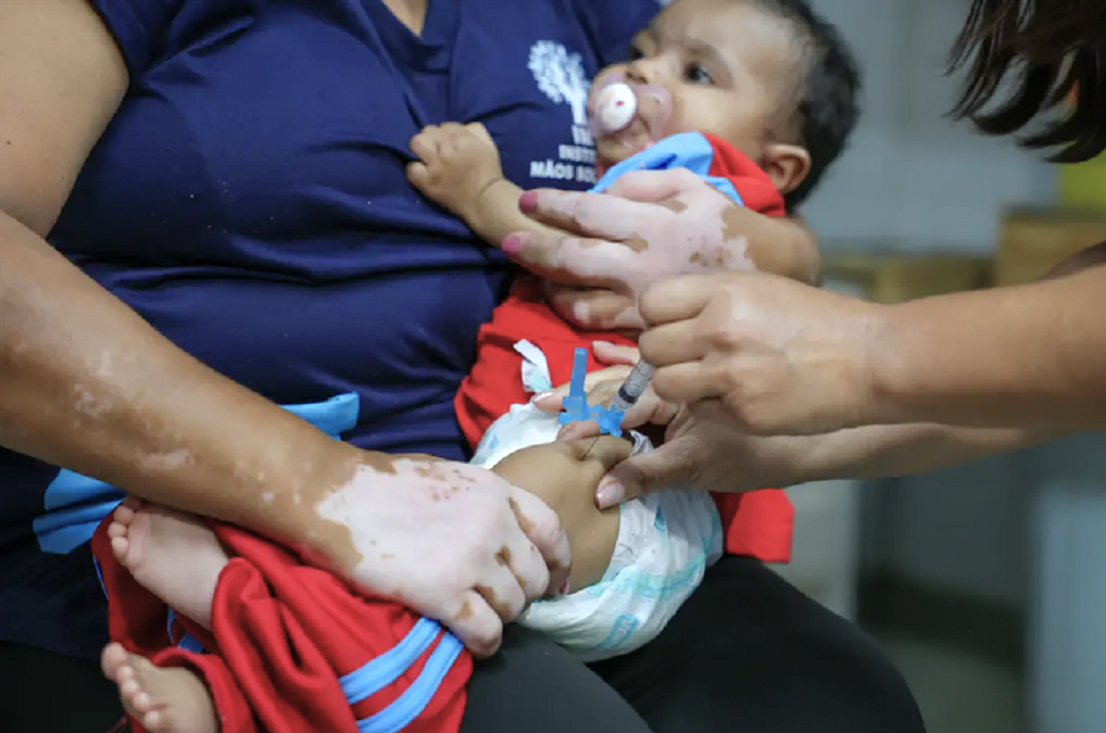 Uma criança é segurada no colo por um adulto enquanto um profissional de saúde aplica uma vacina na coxa. A imagem ilustra a imunização infantil e acompanha reportagem sobre a redução do número de crianças sem a primeira dose da vacina pentavalente no Brasil, segundo dados da OMS e do Unicef.