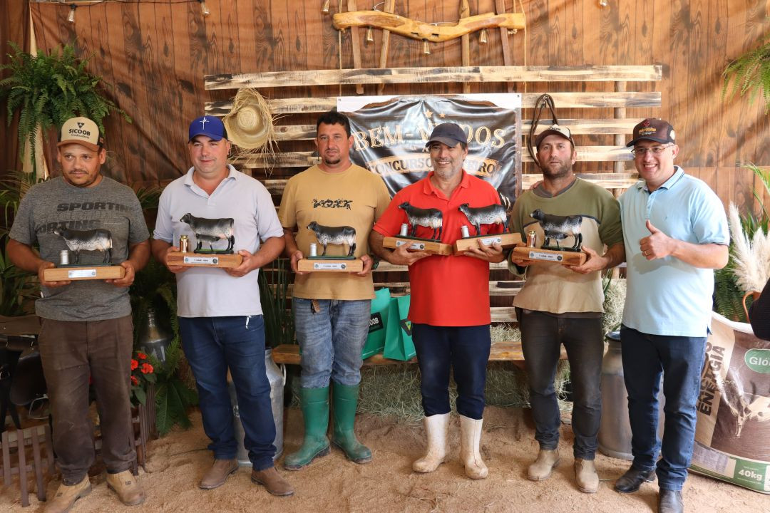 Seis homens alinhados em um ambiente decorado com feno e madeira, segurando troféus de madeira em formato de vacas leiteiras. Alguns usam bonés e botas.