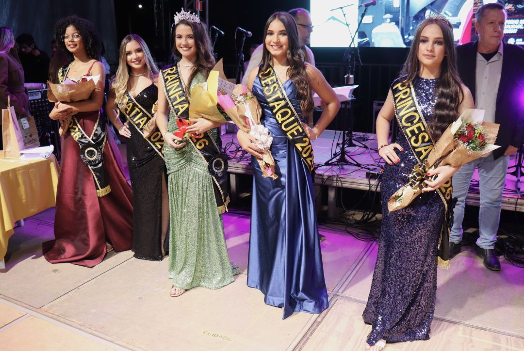 Cinco jovens posam no palco vestindo vestidos de festa em cores variadas, portando faixas de premiação como Rainha, Princesa e Simpatia, além de buquês de flores.