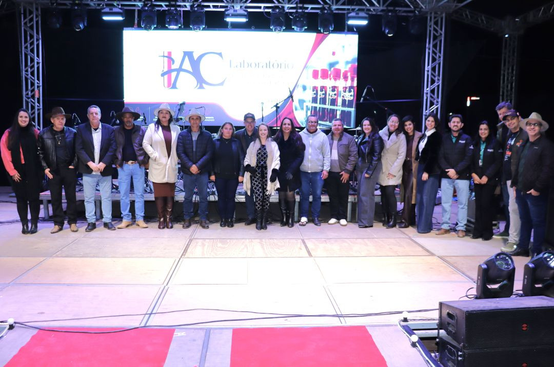 Grupo de homens e mulheres alinhados no palco diante de um painel de LED. A maioria veste trajes de frio e chapéus.