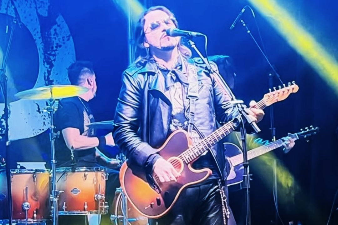 Homem de cabelos compridos e óculos escuros vestindo jaqueta preta de couro, cantando ao microfone e tocando violão no palco sob iluminação azul e amarela.