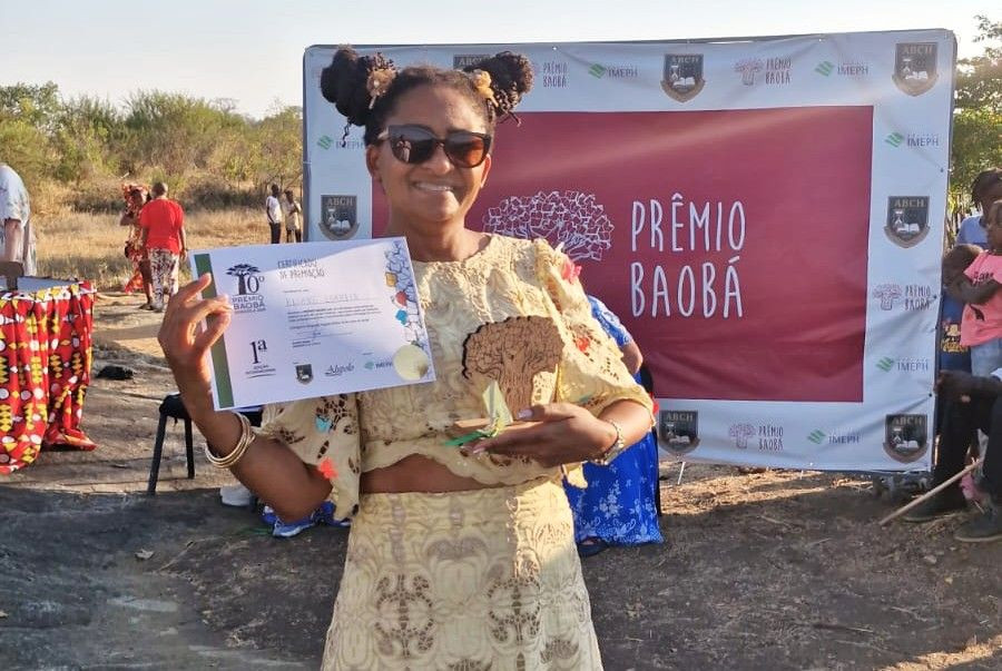 Eliane Correia sorri ao ar livre enquanto segura o certificado de premiação e o troféu do 10º Prêmio Baobá, em frente ao painel do evento em Angola.
