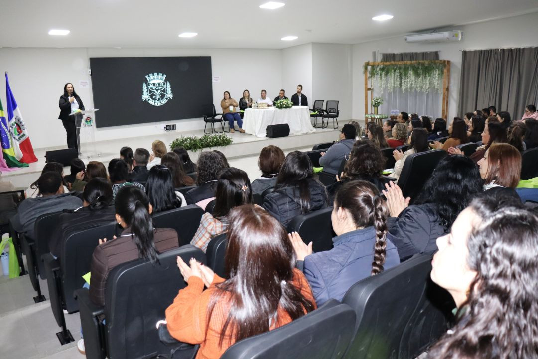 Fotografia colorida tirada da parte de trás de um auditório lotado. Dezenas de educadores estão sentados em poltronas pretas, de costas para a câmera, assistindo à cerimônia no palco ao fundo. No palco, uma mulher vestindo preto fala ao microfone em um púlpito à esquerda. Ao centro do palco, há uma mesa com autoridade sentadas e um grande painel de LED exibindo o brasão do município em verde. Bandeiras do Brasil e do município estão hasteadas à esquerda. A iluminação é embutida no teto branco.