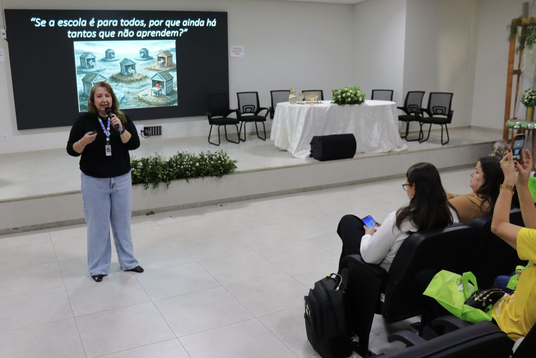 Fotografia colorida em plano médio mostrando uma mulher de cabelos claros, vestindo blusa preta e calça jeans clara, em pé no palco de um auditório. Ela segura um microfone e fala. Atrás dela, um grande telão exibe uma imagem ilustrativa de várias pequenas casas de madeira em um terreno acinzentado, com a frase em branco no topo: “Se a escola é para todos, por que ainda há tantos que não aprendem?”. Na plateia, em primeiro plano à direita, algumas pessoas assistem, e uma mulher levanta o celular para tirar uma foto da palestrante. Há cadeiras vazias na mesa de honra ao fundo do palco.