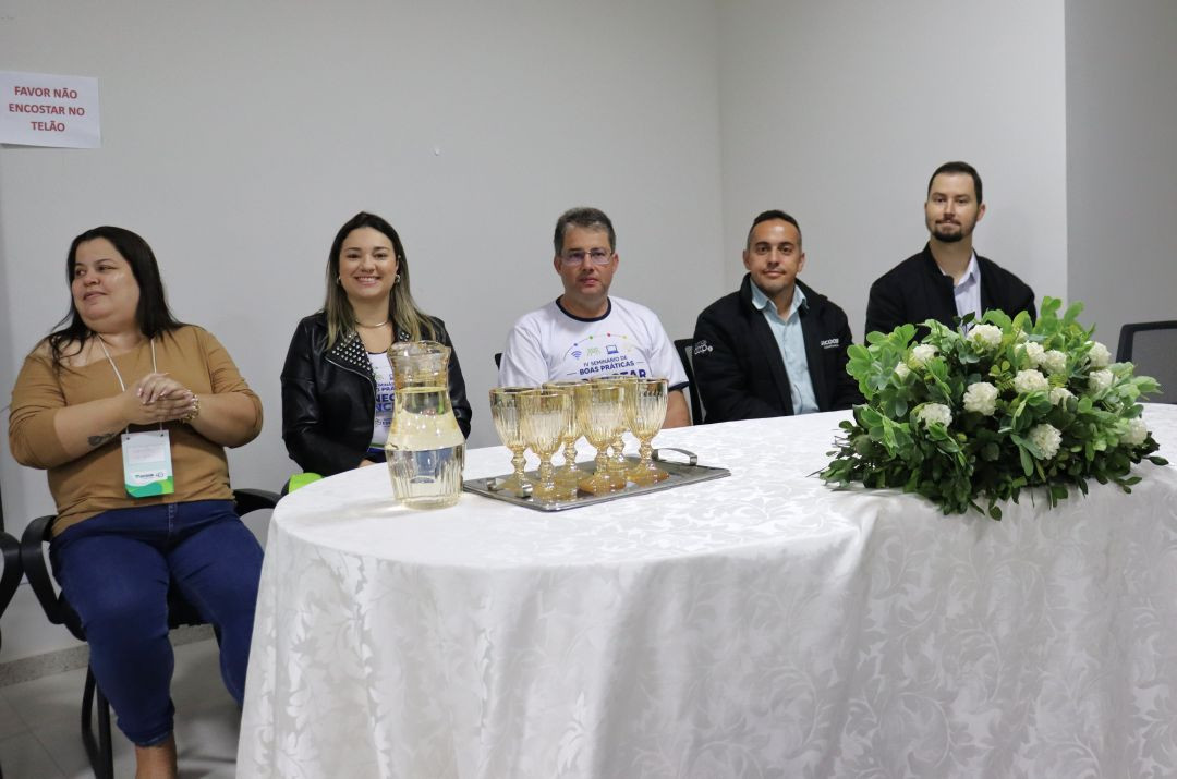 Fotografia colorida em plano médio de cinco pessoas sentadas atrás de uma mesa de conferência coberta com uma toalha branca rendada. Eles estão voltados para a frente, com expressões sérias e respeitosas (posição de hino). Da esquerda para a direita: uma mulher de blusa caramelo e calça jeans; uma mulher de jaqueta de couro preta; um homem de camiseta branca com o logo do evento; um homem de jaqueta preta com logo do Sicoob; e um homem de camisa social branca e paletó preto. Sobre a mesa, há uma bandeja de prata com taças douradas, uma jarra de água e um arranjo de flores brancas e verdes. Na parede branca ao fundo, um pequeno aviso impresso diz: 