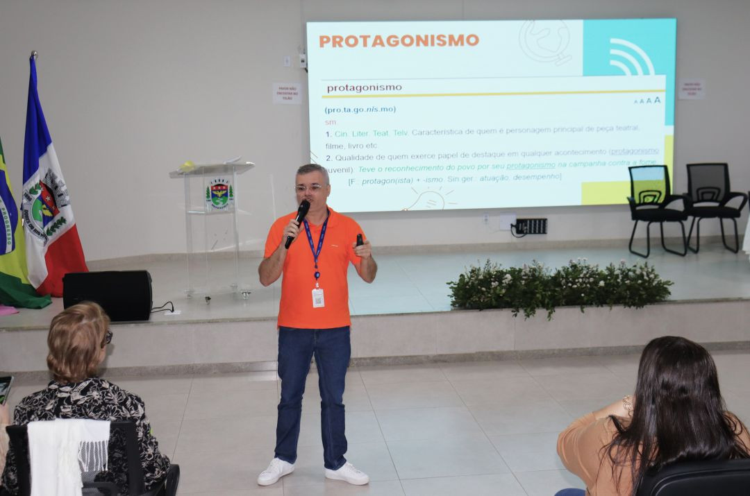 Fotografia colorida em plano médio de um homem de óculos e camisa polo laranja em pé no centro do palco de um auditório. Ele segura um microfone na mão direita e um passador de slides na esquerda, falando para a plateia (da qual se vê apenas as costas de duas pessoas em primeiro plano). Atrás dele, uma grande tela de projeção exibe a palavra 