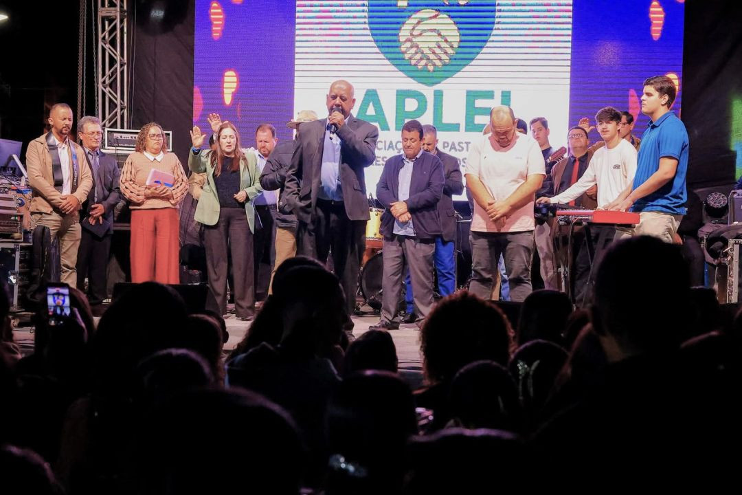 Grupo de líderes religiosos no palco principal com microfone e mãos levantadas durante oração.