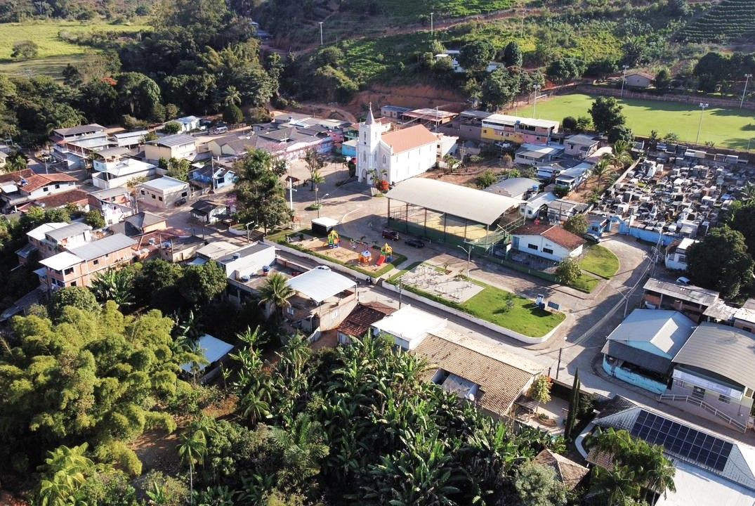 Fotografia aérea panorâmica do distrito de São Tiago, mostrando a igreja matriz no centro, a praça principal, quadra coberta, campo de futebol e casas ao redor.