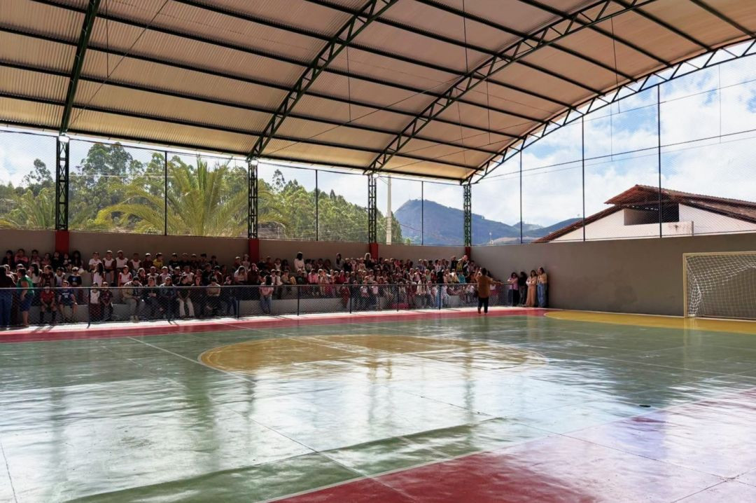 Vista ampla da quadra poliesportiva coberta com estrutura metálica, com plateia sentada na arquibancada lateral e morros ao fundo.