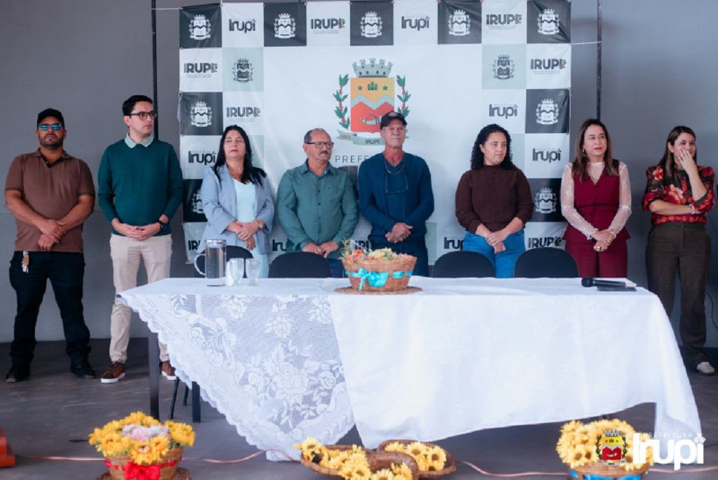 Autoridades e gestores públicos perfilados atrás de uma mesa coberta por toalha branca decorada com arranjos de flores amarelas.