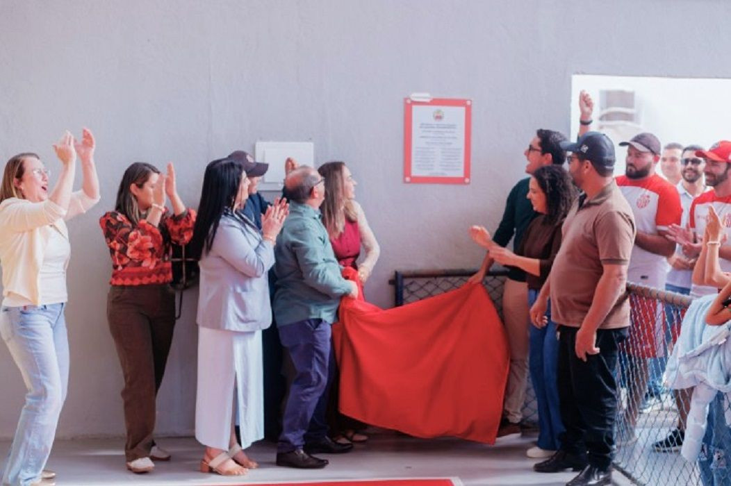 Grupo de pessoas reunidas ao lado de uma placa com moldura vermelha fixada na parede, celebrando o descerramento de um tecido vermelho em solenidade oficial.