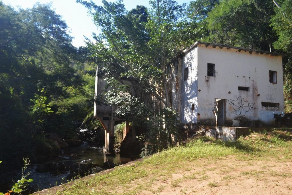 Construção antiga e abandonada de alvenaria branca ao lado de um rio e cercada por árvores, com pichações na fachada e estrutura suspensa sobre o leito da água.