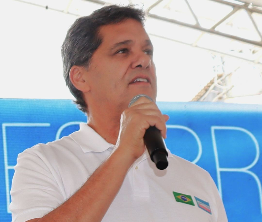 Governador Ricardo Ferraço fala ao microfone em evento do Governo do Espírito Santo, usando camisa branca e tendo, ao fundo, um painel azul com letras brancas e estrutura metálica de uma cobertura.