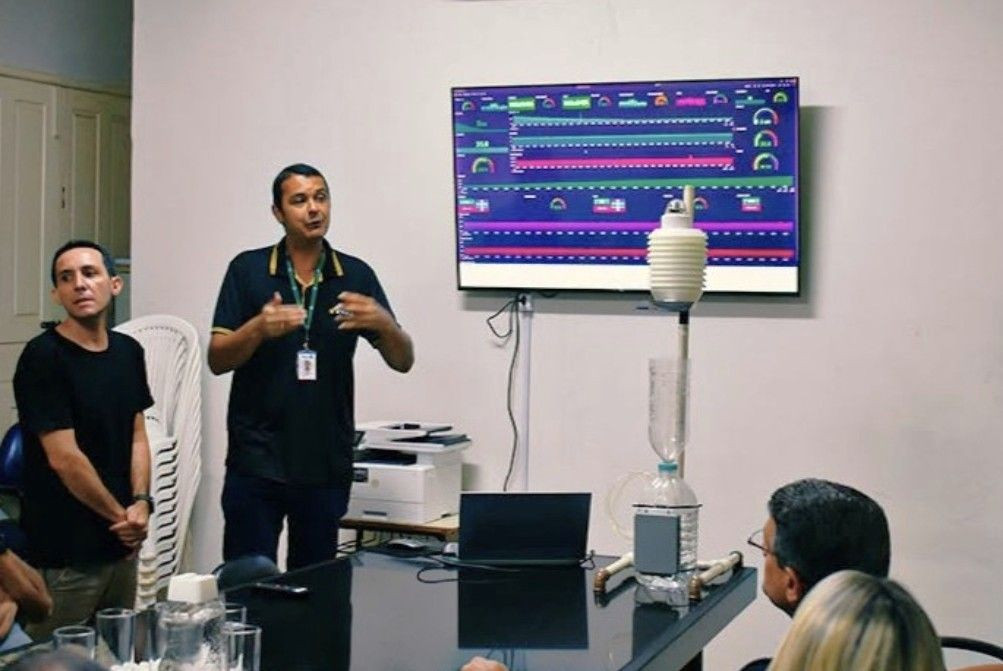 Apresentação do Sistema AlertaES em uma sala de reuniões. Dois homens estão em pé ao lado de um protótipo de monitoramento e de uma tela que exibe gráficos e dados do sistema.