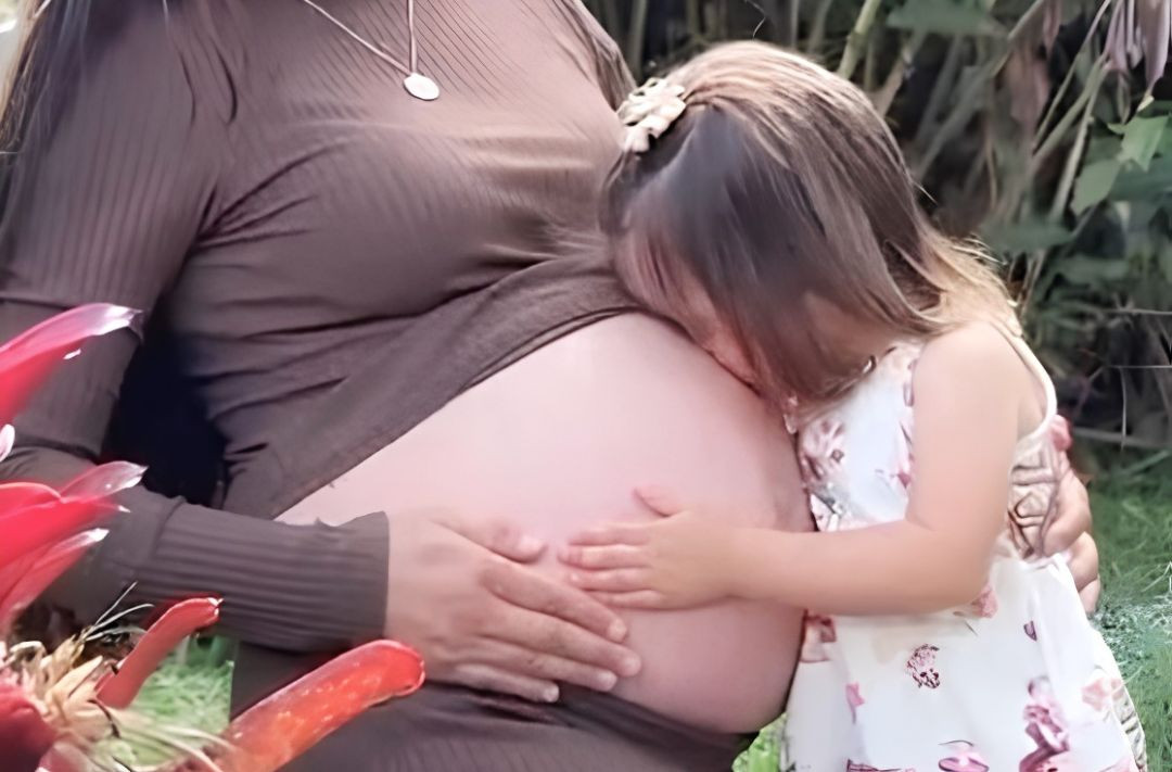 Foto em plano detalhado de uma criança pequena encostando o rosto e abraçando a barriga de uma mulher grávida vestida com blusa marrom.
