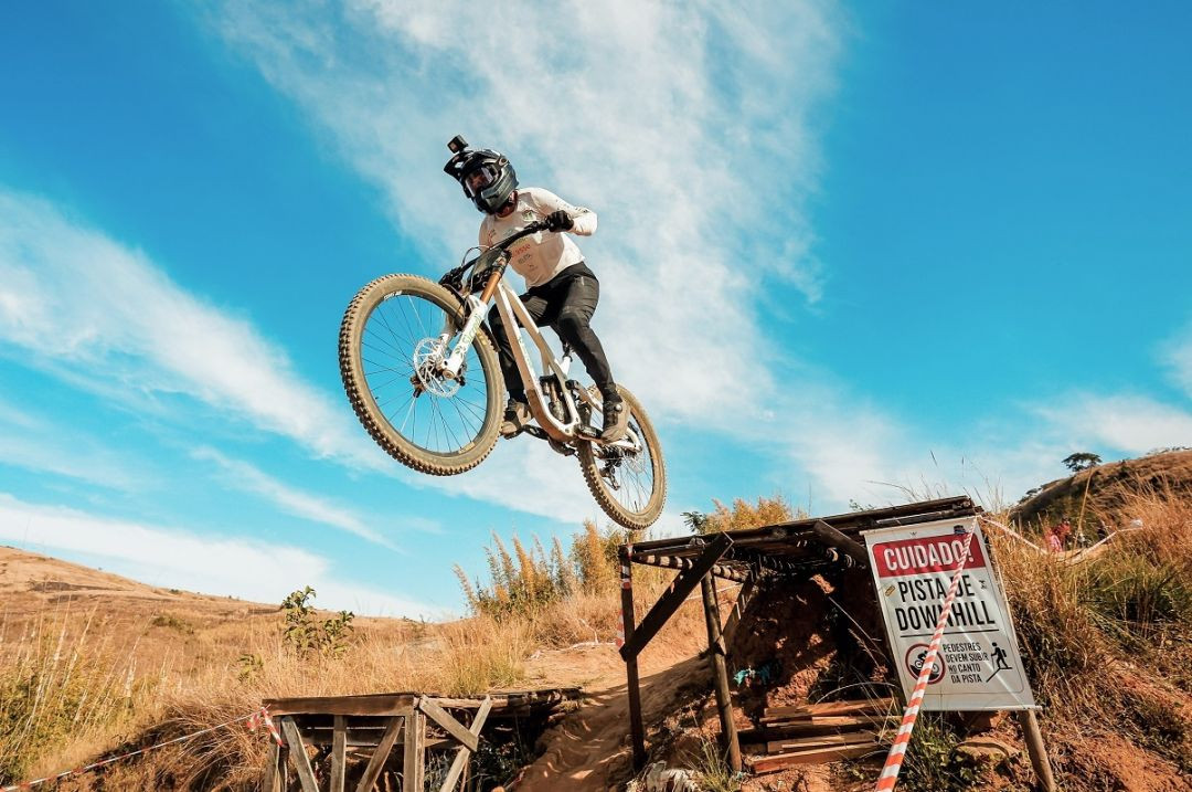 Ciclista no ar executando um salto sobre uma rampa de madeira em pista de downhill. Ao fundo, céu azul com poucas nuvens e morro seco com placa de sinalização 'Cuidado! Pista de Downhill'.