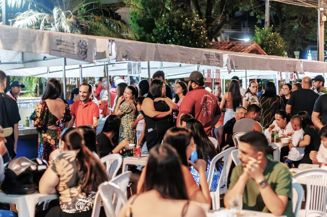 Fotografia noturna da Praça Central de Irupi lotada de pessoas sentadas em mesas plásticas brancas e circulando em frente às barracas da Feira Gastronômica.