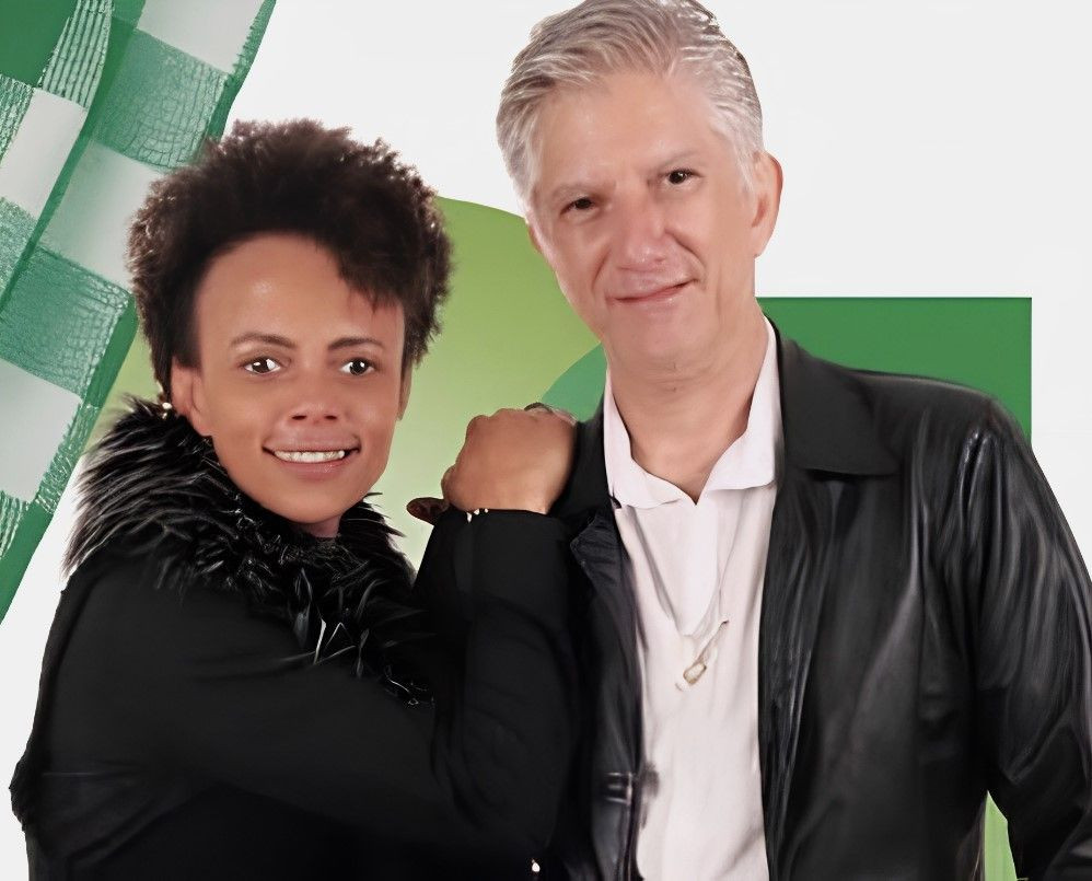 Foto de mulher negra, com cabelos cacheados curtos, usando uma blusa preta com plumas na gola, e um homem branco de cabelo grisalho, com camisa branca e jaqueta de couro preta, que compõem do Trio Comodor's.