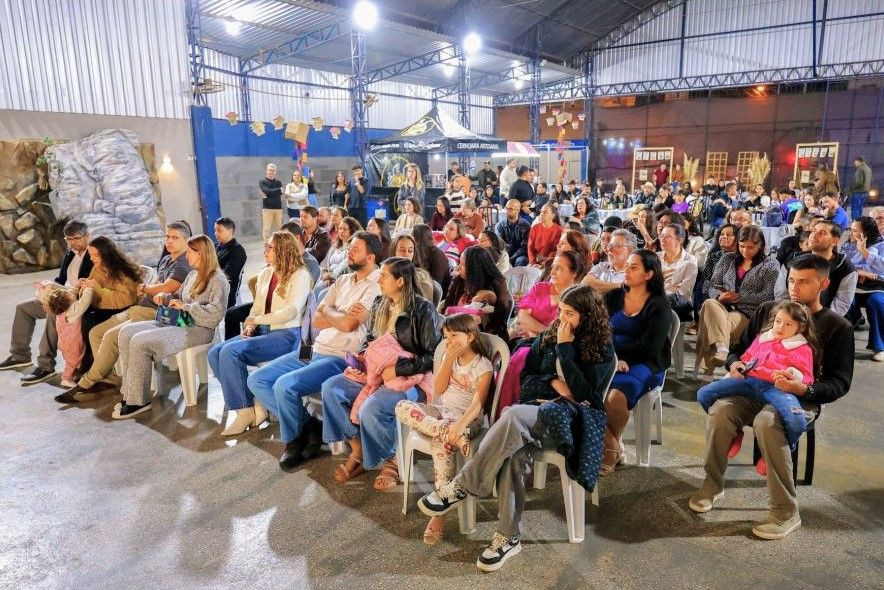 Diversas pessoas de várias idades sentadas em cadeiras brancas de plástico assistindo ao evento em uma quadra coberta.