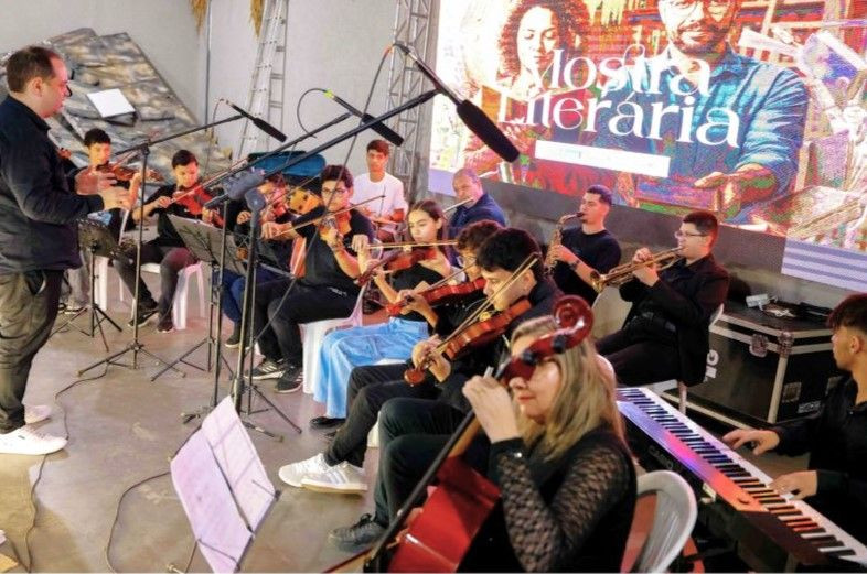 Músicos jovens e adultos tocando violino, violoncelo, teclado e instrumentos de sopro com partituras sob a regência de um maestro.
