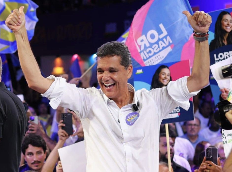 Ricardo Ferraço sorrindo com os braços levantados e fazendo sinal de 'jóia', vestindo camisa branca com adesivo de campanha, cercado por apoiadores com bandeiras ao fundo.