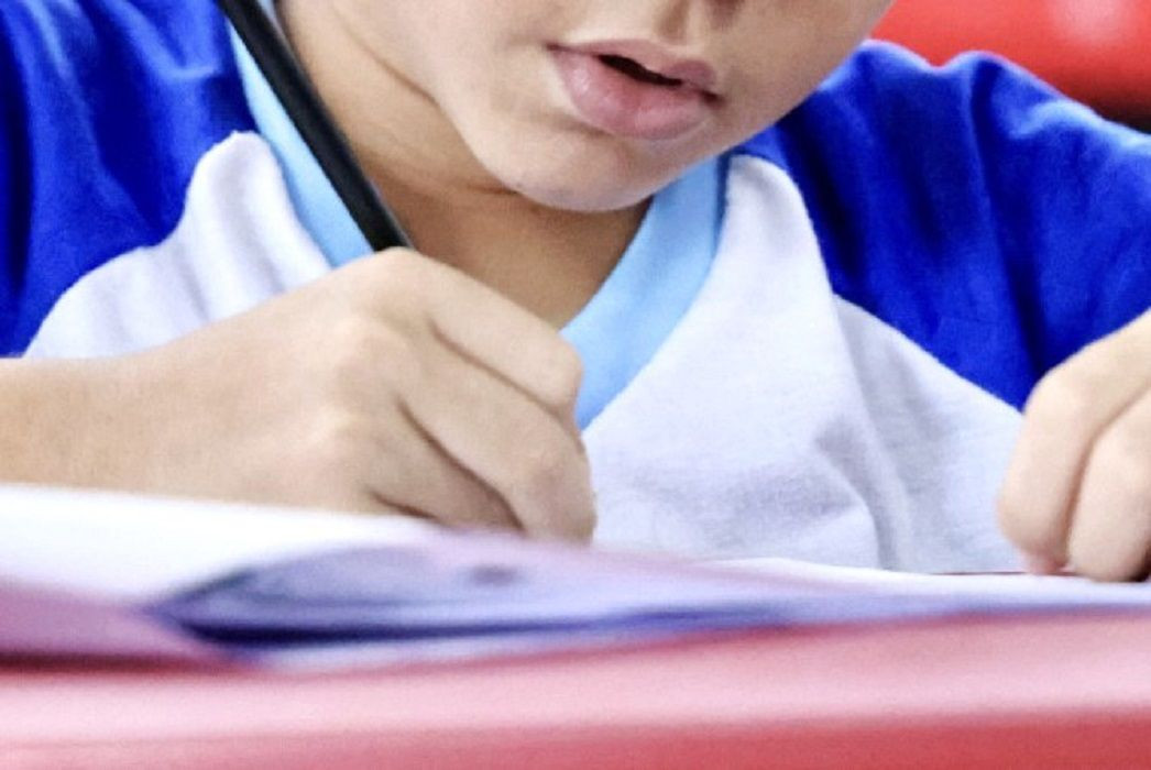 Foto em close mostra um estudante escrevendo em um caderno durante atividade escolar, com foco nas mãos, no lápis e parte do uniforme, ilustrando o tema da educação e do Ideb.
