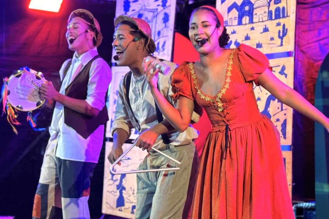 Três atores caracterizados no palco, sendo dois homens tocando pandeiro e triângulo e uma mulher vestida de vermelho cantando.