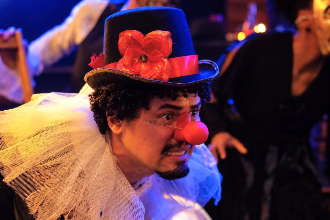 Ator caracterizado de palhaço com nariz vermelho, gola de filó branca e chapéu com flor em destaque no palco.