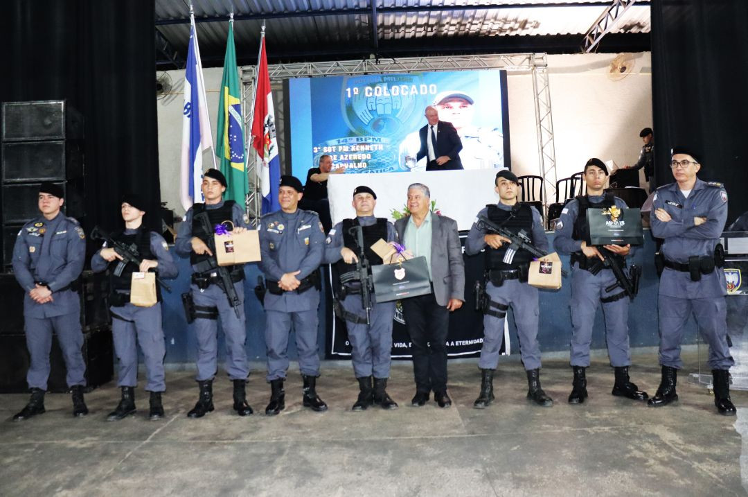 Oito policiais militares e o prefeito Romário Vieira (de terno cinza, ao centro) em pé no palco, à frente da mesa de honra. Os policiais estão de farda cinza e boina preta; alguns seguram fuzis, outros seguram sacolas de presente pretas e pardas. No telão ao fundo, lê-se 