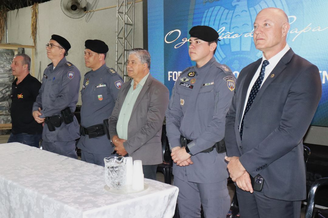 Seis homens em pé atrás de uma mesa coberta com toalha branca rendada. Da esquerda para a direita: um homem de camisa preta com logo da Força Tática; o tenente-coronel Weden Carlos em farda cinza e boina; outro oficial em farda semelhante; o prefeito de Iúna, Romário Vieira, de terno cinza e camisa clara; o major PM Lucas Muzi em farda cinza; e o deputado estadual Coronel Weliton Pereira de terno azul escuro e gravata. No fundo, o telão azul.