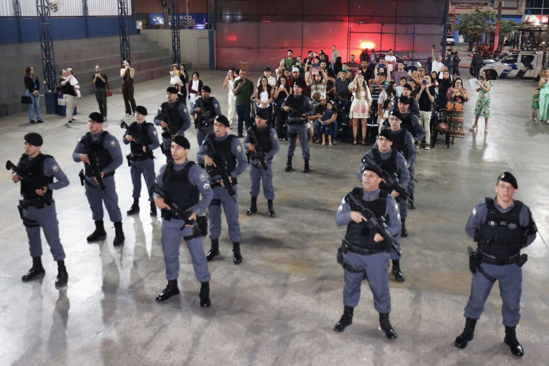 Fotografia em ângulo elevado mostrando o salão do Centro Multiuso. No centro, doze policiais militares da Força Tática em farda cinza e com fuzis estão perfilados em duas fileiras. Atrás deles, uma plateia de civis (familiares e convidados) está em pé observando. À direita, um carro de polícia branco e azul está estacionado. O ambiente tem piso de cimento e estrutura metálica.