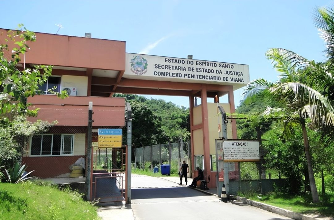 Entrada do Complexo Penitenciário de Viana no Espírito Santo, mostrando o pórtico principal de cor alaranjada com a placa da Secretaria de Estado da Justiça e a portaria de acesso com agentes de segurança.