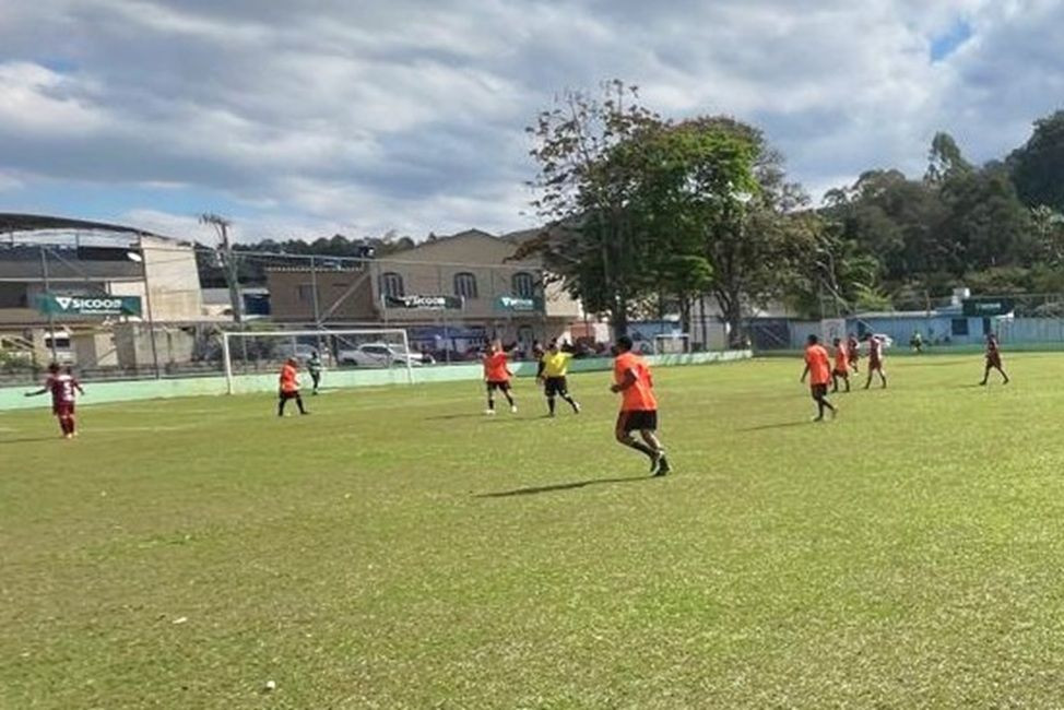 Partida de futebol amador em campo de grama verde em Dores do Rio Preto, sob dia ensolarado. Jogadores vestem uniformes nas cores laranja e vinho, enquanto o árbitro atua no centro do gramado. Ao fundo, veem-se traves, placas publicitárias e residências.