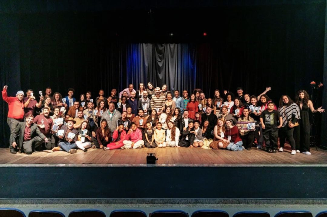 Grupo extenso de artistas e organizadores sorrindo e segurando troféus no palco de um teatro com cortina escura ao fundo.