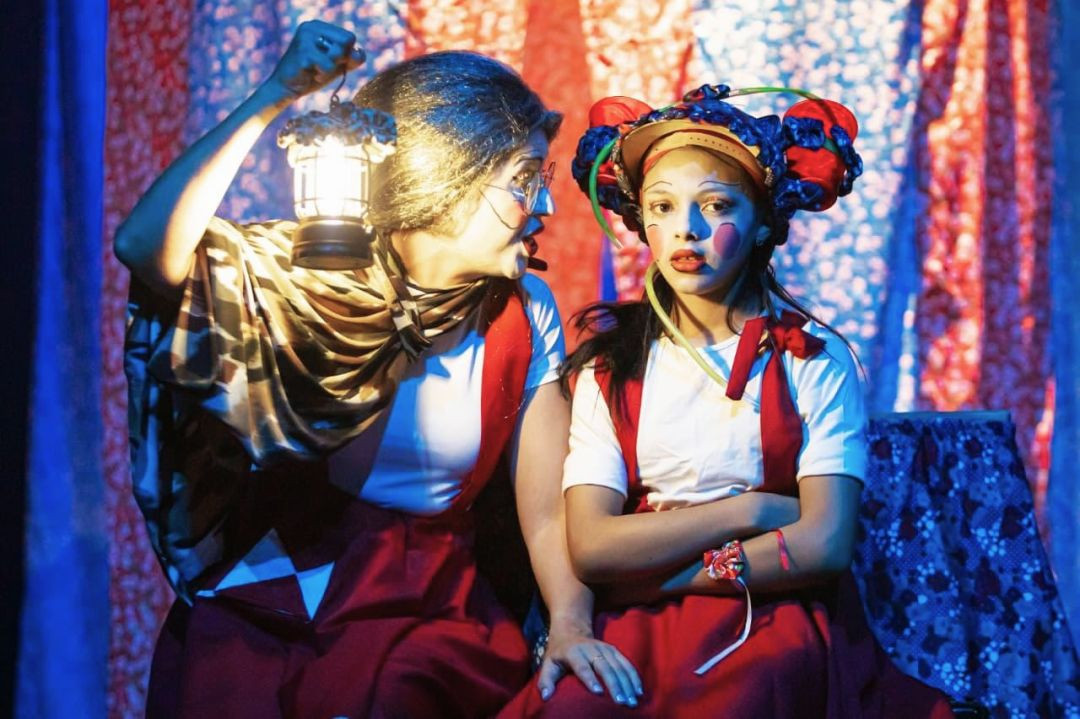 Duas atrizes caracterizadas no palco; uma senhora segurando uma luminária acesa ao lado de uma jovem maquiada com adereços coloridos na cabeça.